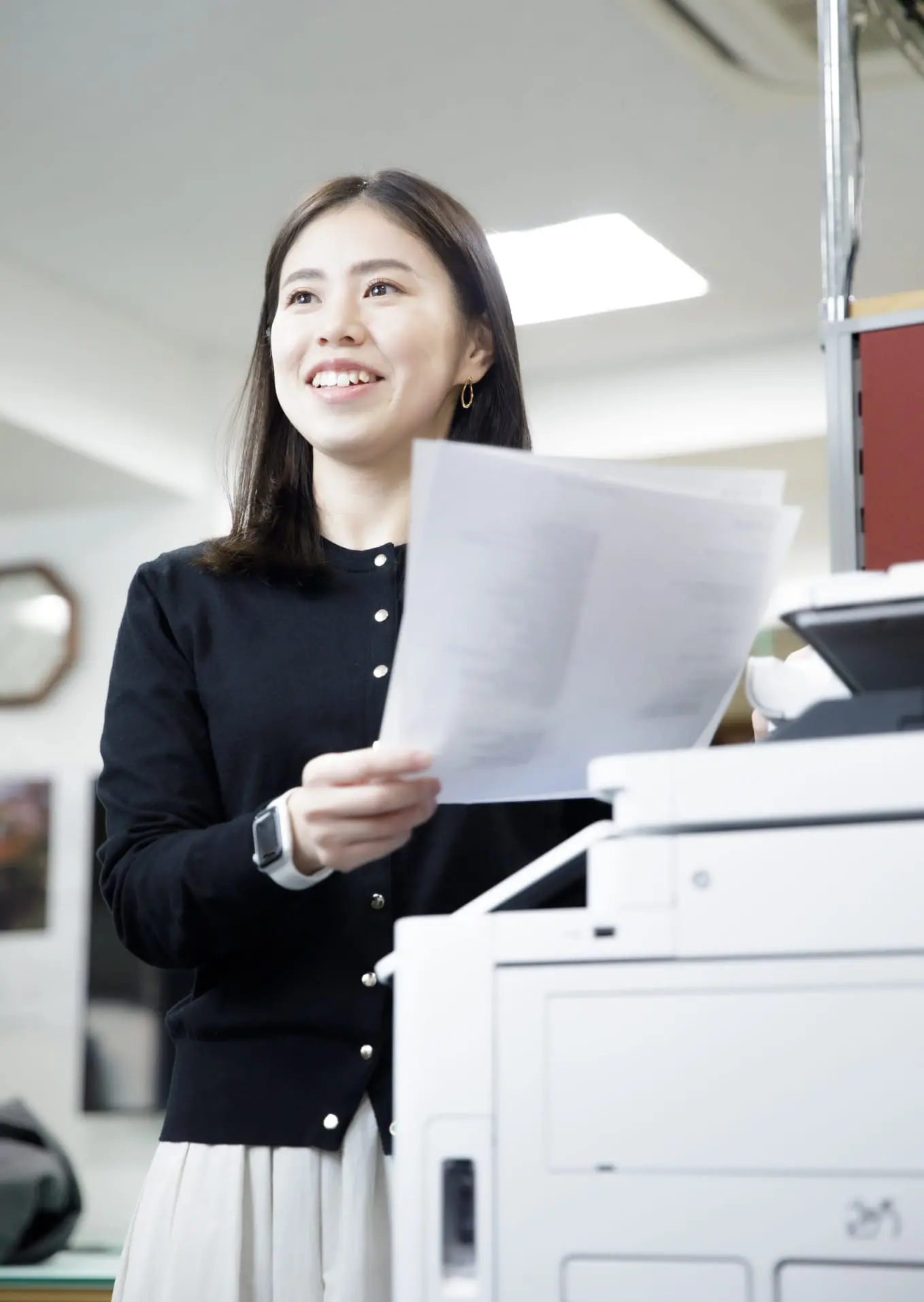 社員の女性がコピー機の前で資料を持ちながら笑顔で立っている様子